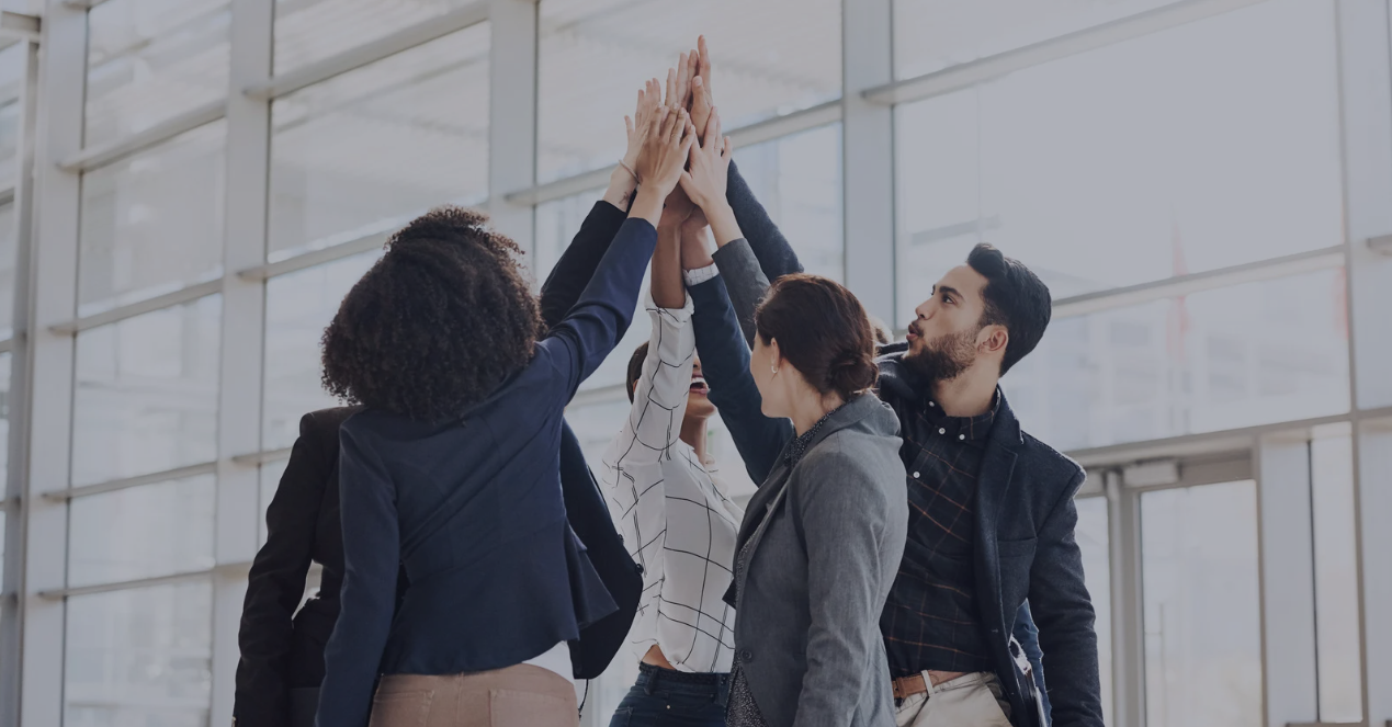 A diverse team of professionals in business attire celebrating with a group high five, symbolizing teamwork, collaboration, and shared mission.