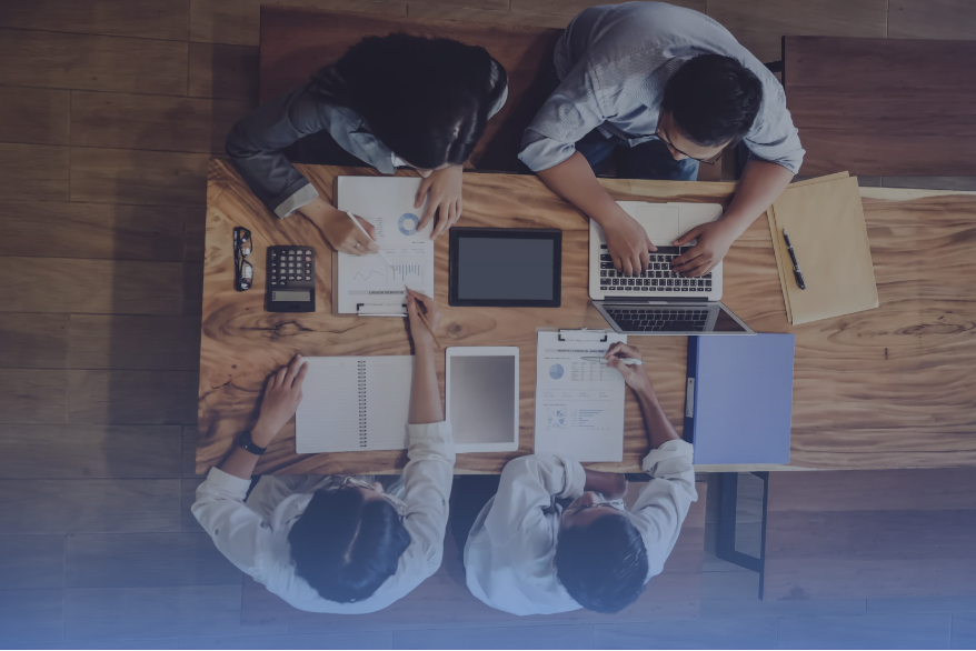 Overhead view of a diverse team working together at a wooden conference table with laptops, documents, and digital tablets.